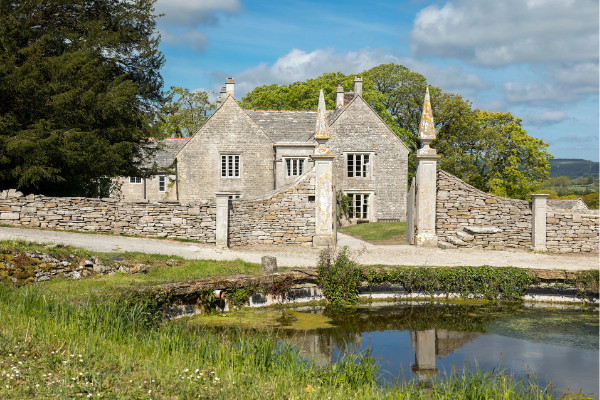 The exterior frontage of Dunshay Manor with a blue sky behind it. A pale grey stoned manor with two spiked stone gate piers. In the foreground is a pond. 