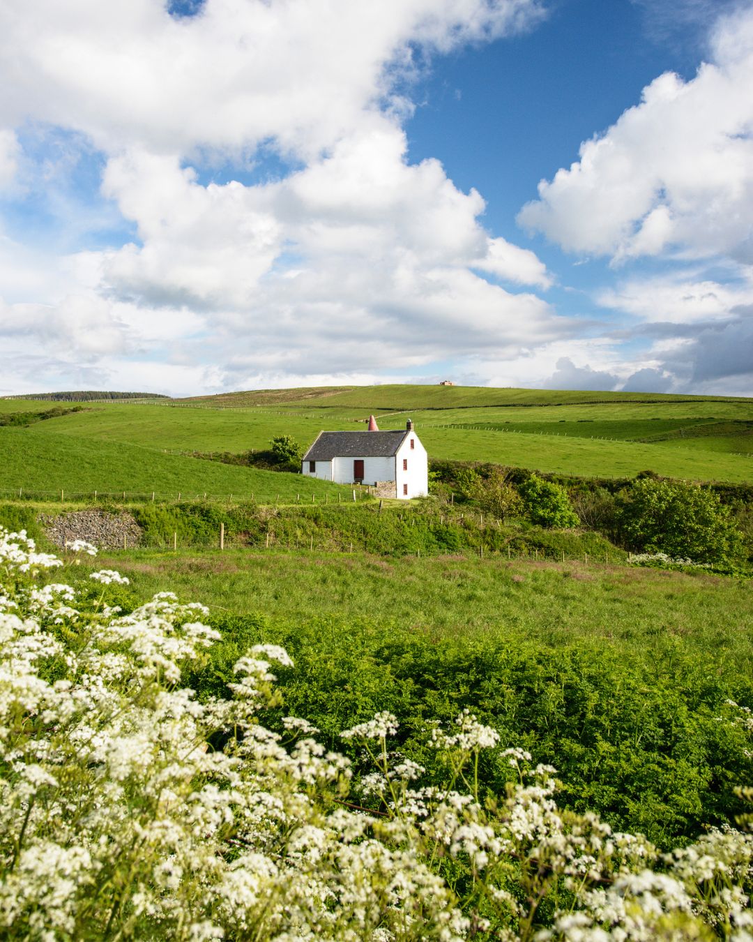 A small white mill building from afar, surrounded by green Scottish landscape